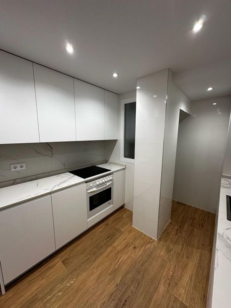 Cocina moderna blanca con suelo de madera, horno empotrado y pasillo estrecho.