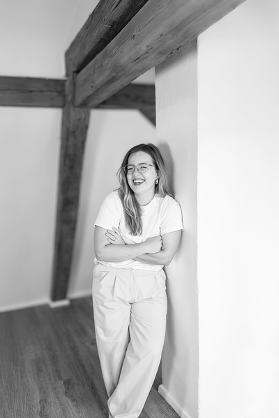Gründerin Lea Kohlhaas, an eine weiße Wand gelehnt, Arme verschränkt.Holzbalken über dem Kopf. Schwarz und weiß.