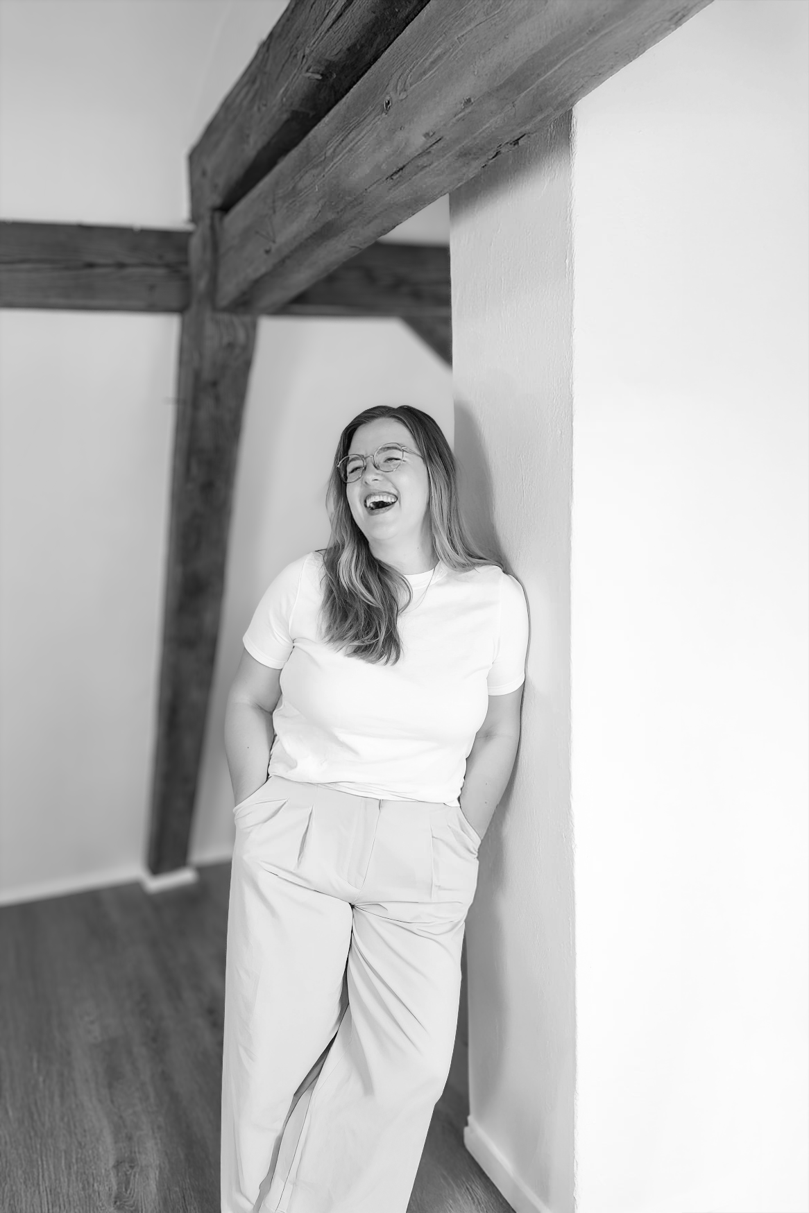 Gründerin Lea Kohlhaas, an eine weiße Wand gelehnt, Hände in den Taschen. Holzbalken über dem Kopf. Schwarz und weiß.