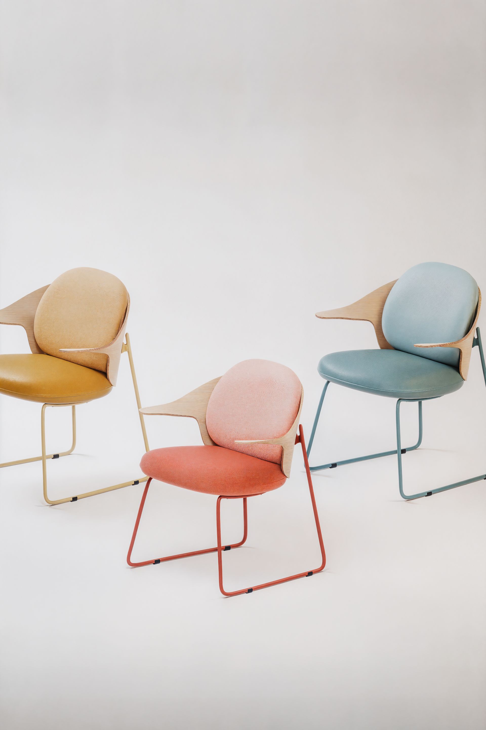 Drei moderne Stühle in Gelb, Rosa und Blau mit Holzarmlehnen vor weißem Hintergrund.