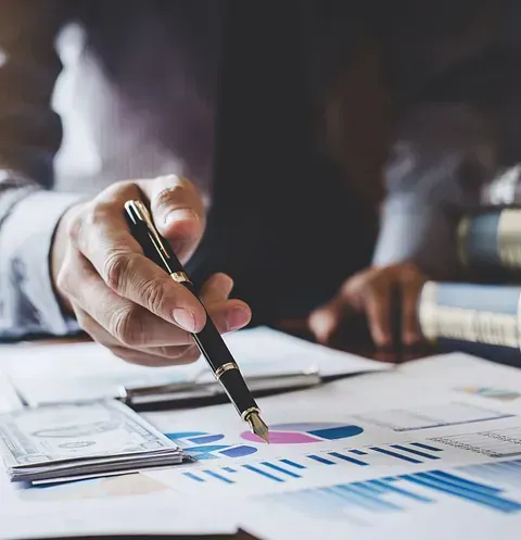 Una persona señala con un bolígrafo un gráfico financiero. Hay dinero cerca.