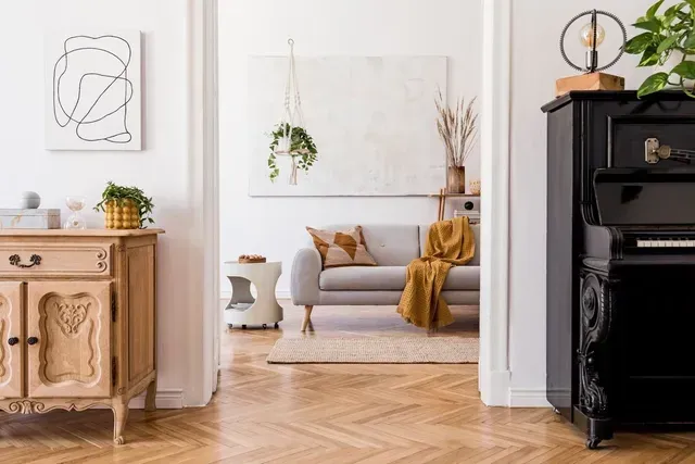 Sala de estar luminosa con sofá beige, manta color mostaza, planta colgante y suelo de madera en espiga.