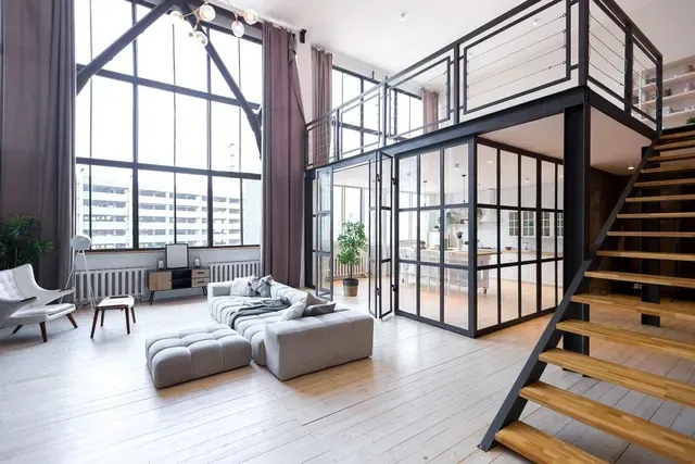 Luminoso salón tipo loft moderno con grandes ventanales, asientos grises y una escalera abierta.