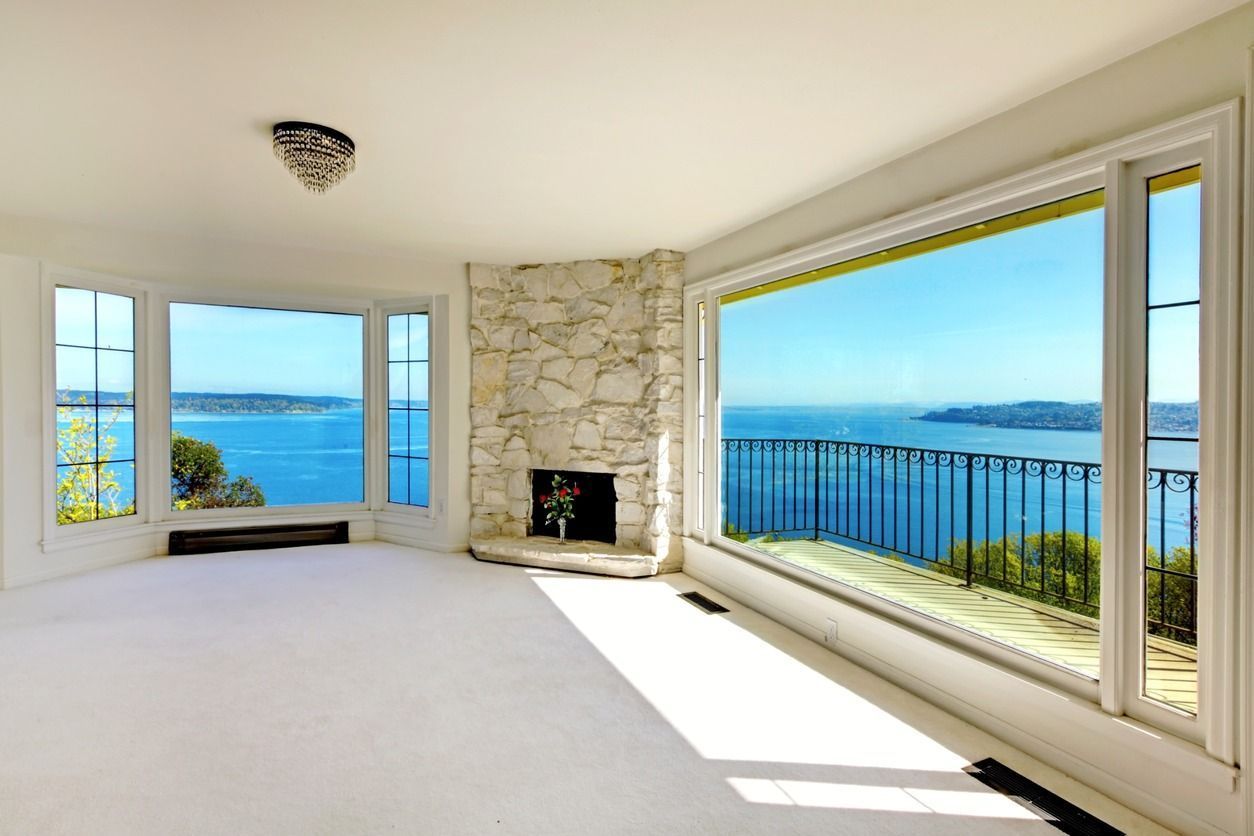 Luminoso salón vacío con chimenea de piedra y grandes ventanales con vistas al océano azul.