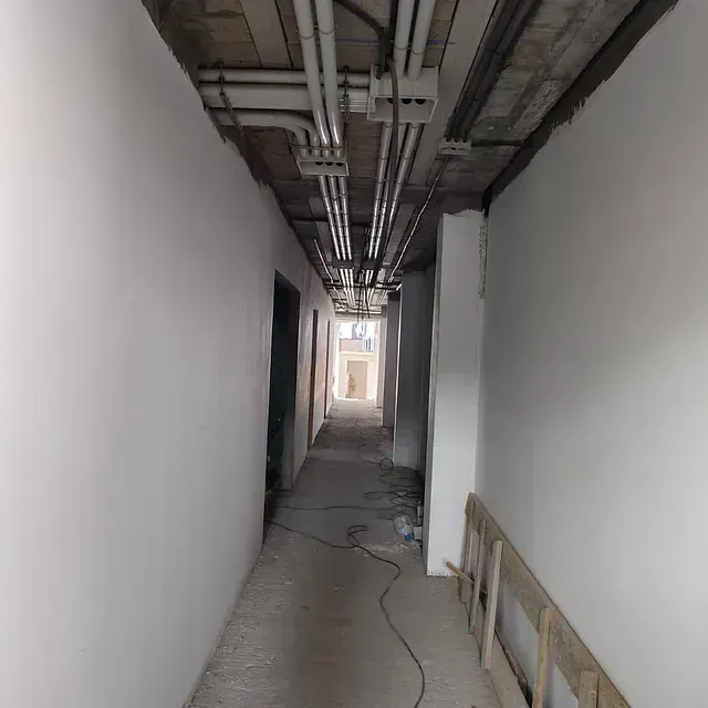 Pasillo estrecho y sin terminar, con tuberías de techo a la vista, paredes blancas y suelo de hormigón con cables.
