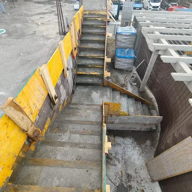 Escalera industrial y zanja de hormigón junto a una barrera de seguridad amarilla en una obra en construcción.