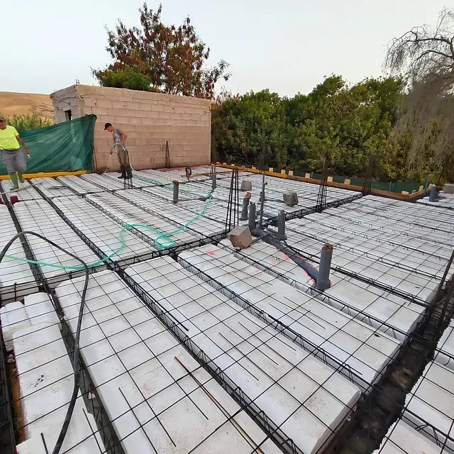 Obra en construcción con rejilla de armadura y trabajadores preparando una cimentación de losa de hormigón.
