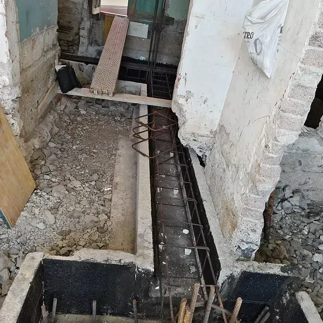 Zanja de construcción con barras de refuerzo expuestas y cimientos de hormigón entre paredes sin terminar.