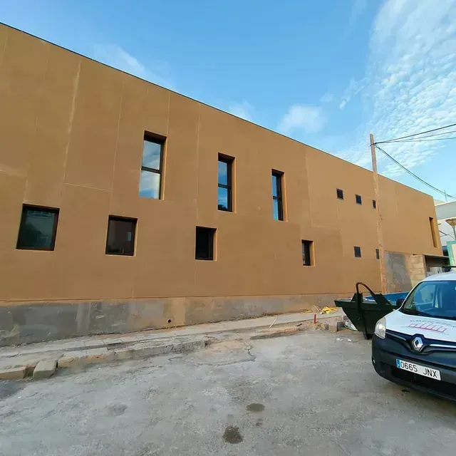 Edificio con fachada de estuco color canela, ventanas estrechas y un coche azul oscuro aparcado delante.