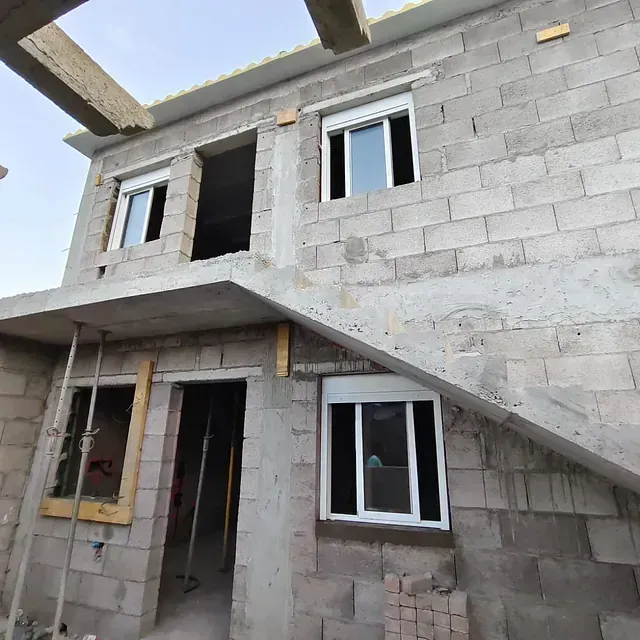 Casa de dos plantas de hormigón sin terminar con paredes de bloques a la vista y ventanas en construcción.