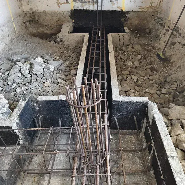 Obra en construcción con cimientos de hormigón, barras de refuerzo expuestas y escombros a lo largo de una zanja central.