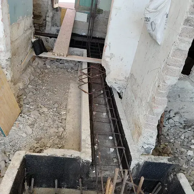 Obra en construcción con zanja de cimentación expuesta, barras de refuerzo y escombros.