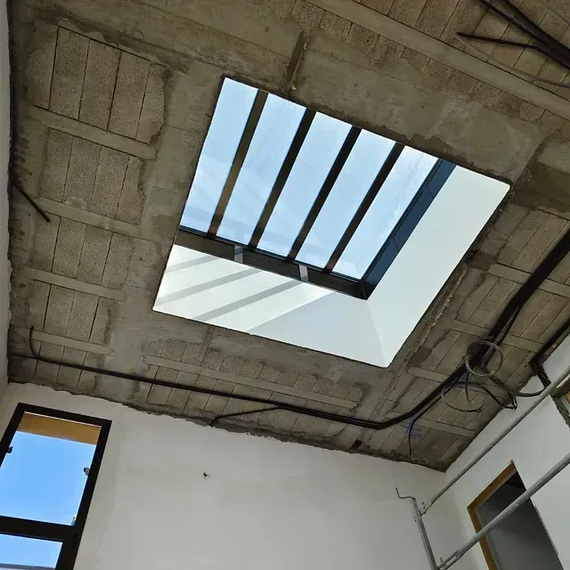 Vista hacia arriba de un techo de hormigón con una claraboya cuadrada y vigas a la vista, sobre una habitación blanca.