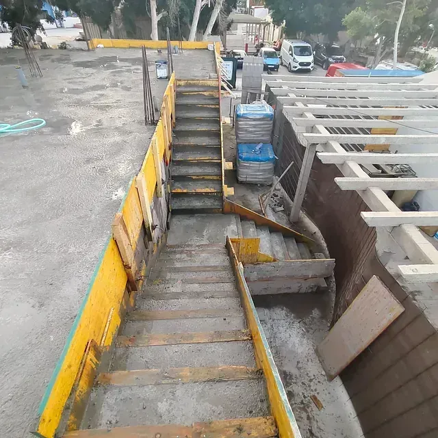Zanja de construcción con muros de seguridad amarillos, escalones de hormigón y materiales apilados junto a una carretera.