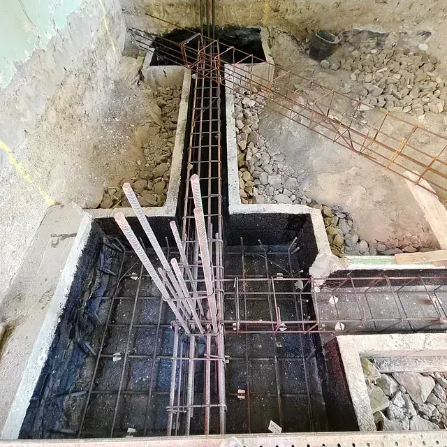 Obra en construcción con cimientos de hormigón y barras de refuerzo expuestas en una zanja revestida de piedra.