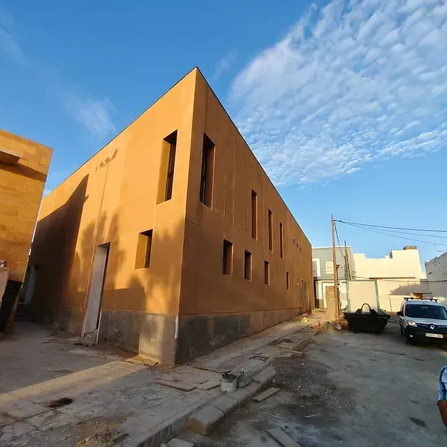 Edificio de color beige bañado por el sol en una calle estrecha bajo un cielo azul, con coches aparcados.