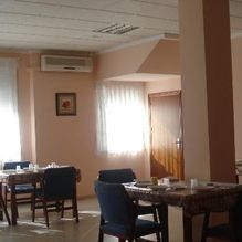 Pequeño comedor con mesas, sillas, aire acondicionado de pared y luz natural que entra por una ventana con cortinas.