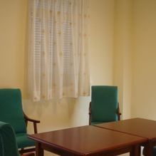 Pequeña sala de reuniones con dos sillas verdes, una mesa de madera y una ventana con cortinas transparentes.