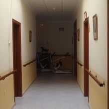 Pasillo de hospital con poca luz, paredes de color beige, pasamanos y una bicicleta estática al final.
