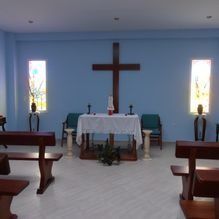 Interior de una pequeña capilla con altar, una gran cruz de madera y bancos orientados hacia el frente.