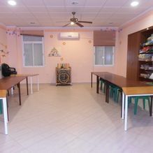 Sala comunitaria vacía con mesas a ambos lados y un altavoz en la pared del fondo, iluminada por luces de techo.