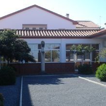 Casa de una sola planta con tejado de tejas rojas, ventanas frontales y entrada para vehículos sombreada bajo la luz del sol.