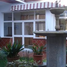 Patio con ventanas de marcos blancos, toldo a rayas, plantas en macetas y una fuente de piedra en la fachada.