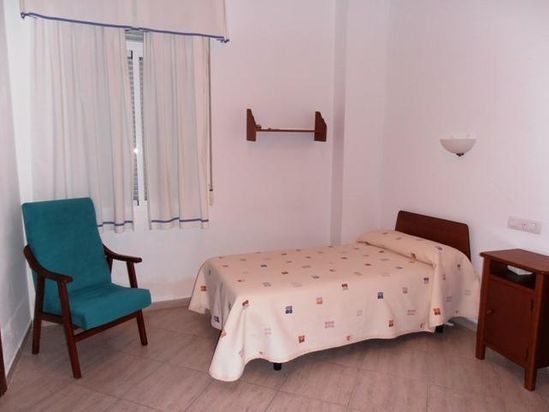 Habitación sencilla con una cama individual, una silla color turquesa, cortinas y una pequeña mesita de noche.