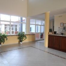 Vestíbulo luminoso con grandes ventanales, plantas en macetas, suelo de baldosas y un mostrador de recepción de madera.