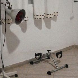 Una pequeña bicicleta estática sobre un suelo de baldosas, junto a paredes blancas y cortinas, con una lámpara roja a la izquierda.