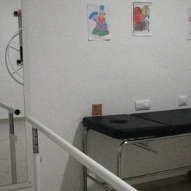 Habitación blanca con una camilla de masaje negra, cuadros en la pared y una barandilla de escalera en primer plano.
