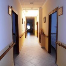 Pasillo largo y luminoso con puertas abiertas, pasamanos de madera y suelo de baldosas en un edificio.