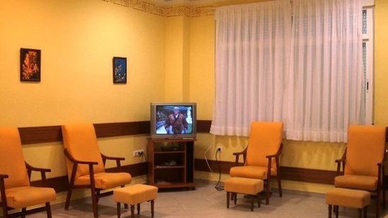 Sala de espera con sillas naranjas, un pequeño televisor y cortinas sobre una gran ventana.