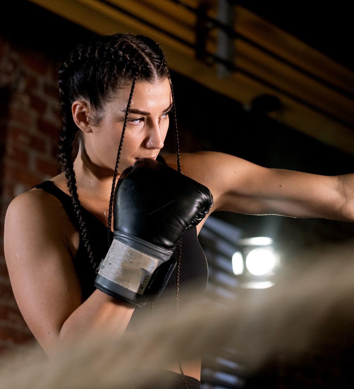Boxeador con camiseta negra sin mangas y guantes, lanzando un puñetazo en un ring de boxeo. Cabello trenzado.