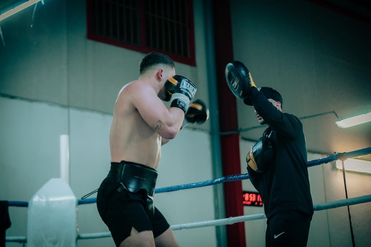 Boxeador con camiseta negra sin mangas y guantes, lanzando un puñetazo en un ring de boxeo. Cabello trenzado.