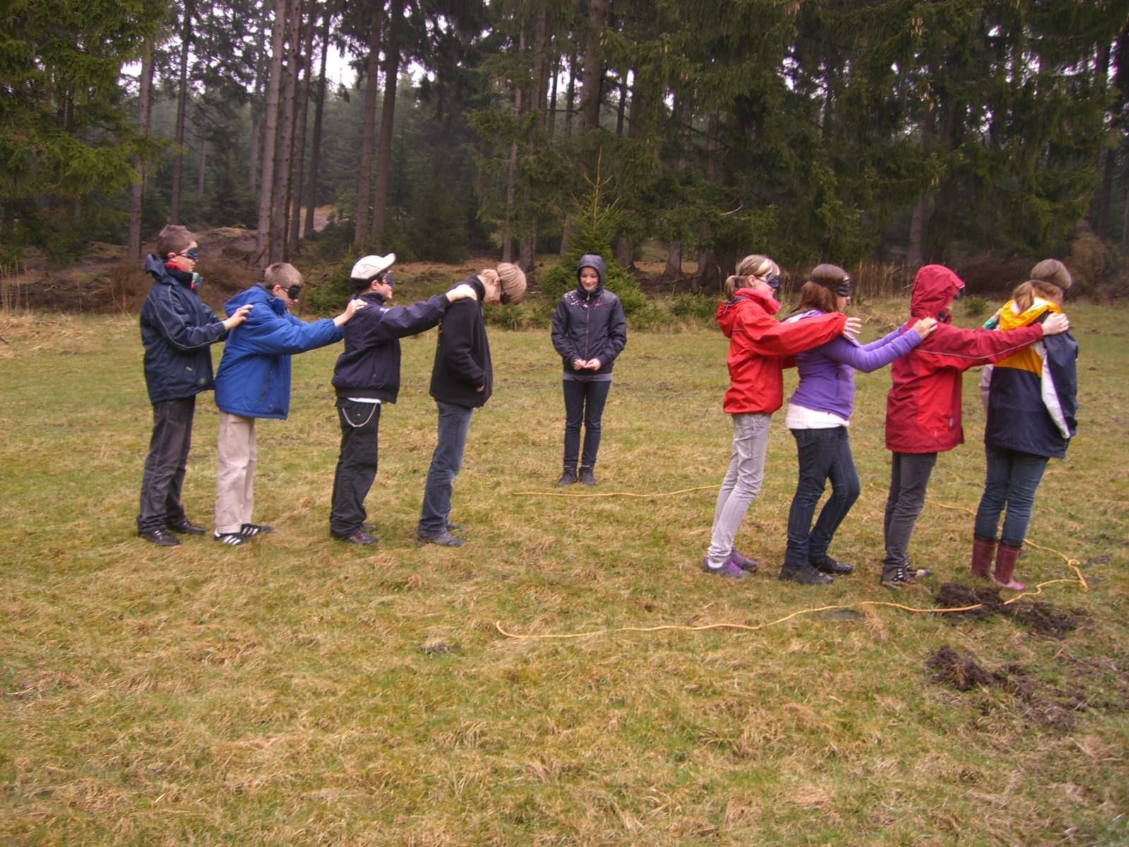 Gruppe von Menschen auf einem Feld, Arm in Arm. Eine Person steht allein. Im Hintergrund Wald.