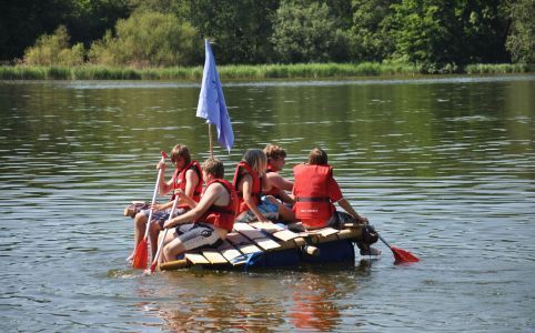 Eine Gruppe von Menschen in Schwimmwesten paddelt auf einem Floß mit blauem Segel auf einem See.