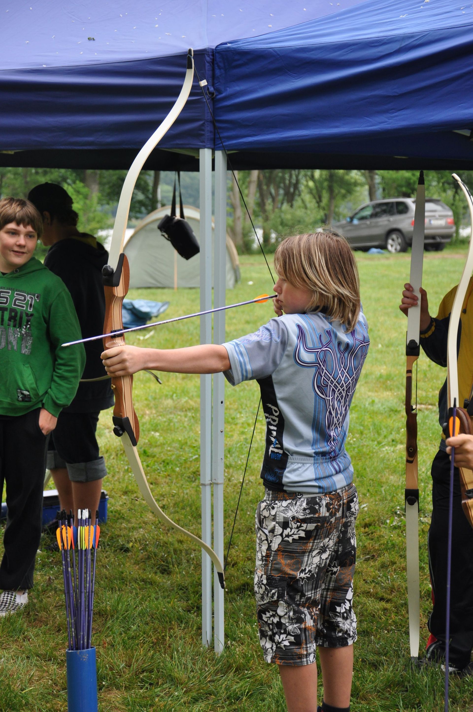 Junger Bogenschütze zielt mit Bogen unter blauem Zelt, im Freien.