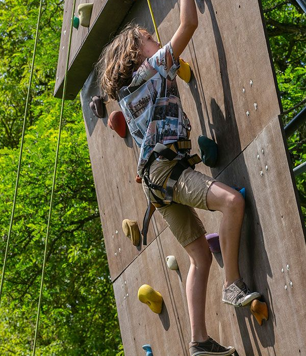 Junge klettert mit einem Klettergurt an einer Felswand im Freien hoch. Er streckt sich nach oben, im Hintergrund ein Wald.