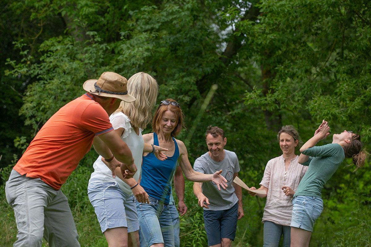 Eine Gruppe von Menschen spielt im Freien ein Spiel und streckt die Arme aus. Im Hintergrund grüne Bäume.