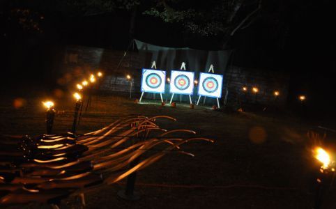 Eine Gruppe von Menschen spielt im Freien ein Spiel und streckt die Arme aus. Im Hintergrund grüne Bäume.