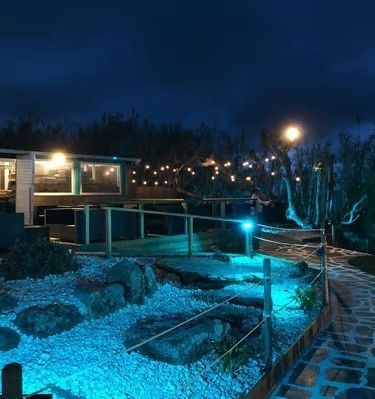 Piscina exterior iluminada con luz azul y jacuzzi por la noche junto a una casa y un sendero de piedra.