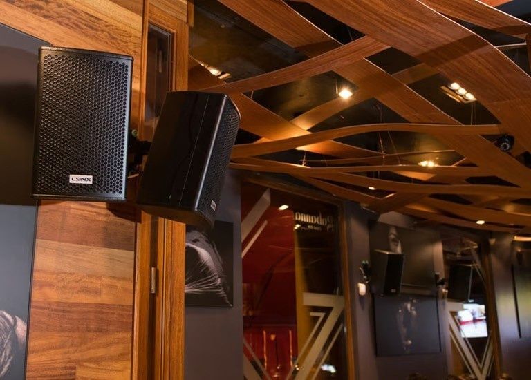 Altavoces de pared negros montados en un techo revestido de madera en el interior de un restaurante o bar.