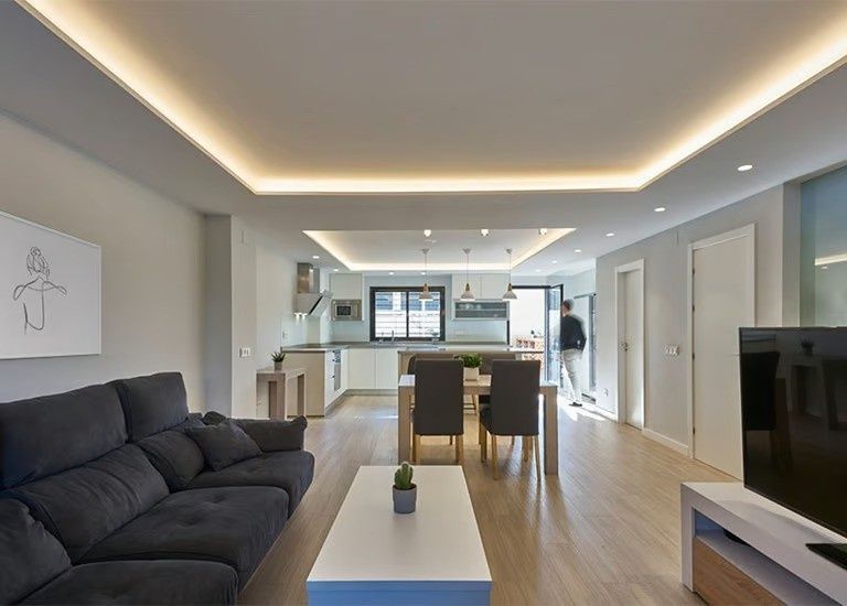 Salón moderno de planta abierta con sofá oscuro, mesa de centro, zona de comedor y cocina con iluminación empotrada.
