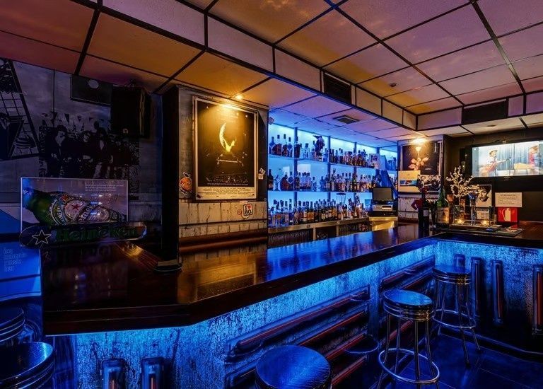 Interior de bar iluminado con luces de neón, con estantes, taburetes y mostrador reflectante retroiluminados