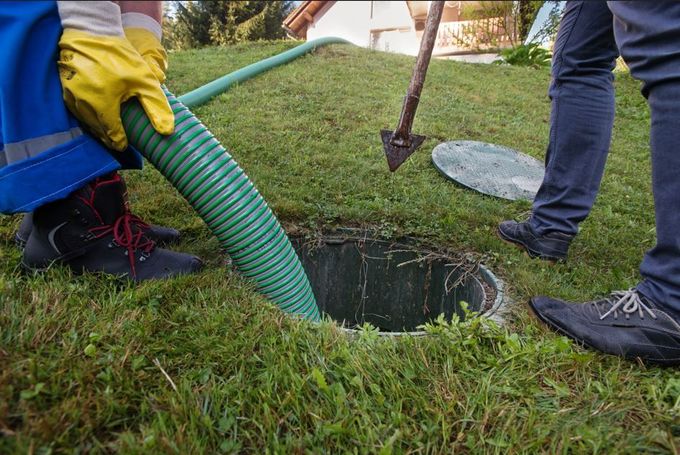 Obreros vaciando un tanque séptico en un césped con una manguera verde, la tapa abierta y una pala.