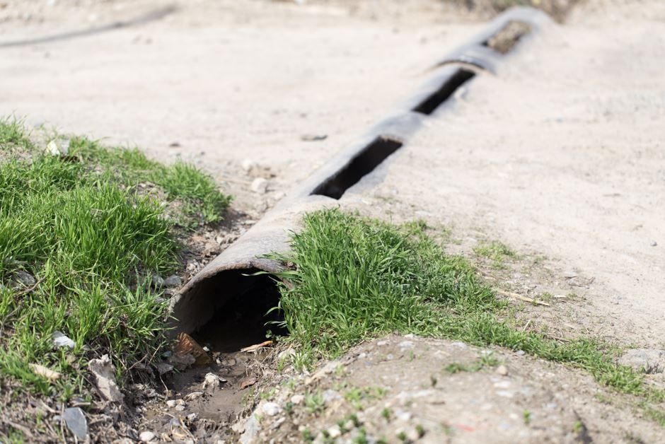 Una alcantarilla de hormigón con aberturas rectangulares se encuentra al borde de un camino de tierra con hierba.