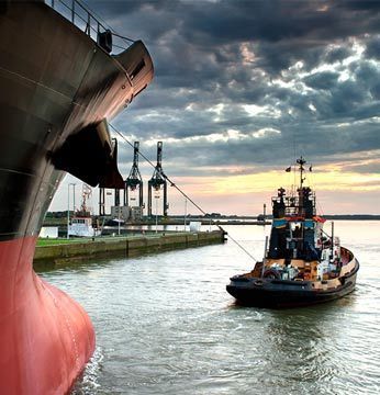 Un remolcador ayuda a un gran buque de carga en un puerto, bajo un cielo nublado.