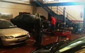 Dos coches en un garaje con una persona con una camisa roja trabajando en un vehículo cerca de una escalera.