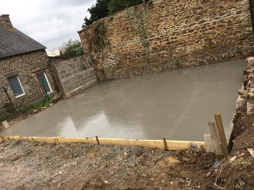 Coulage d'une dalle de béton dans une cour entourée de murs en pierre, à côté d'une maison rustique.